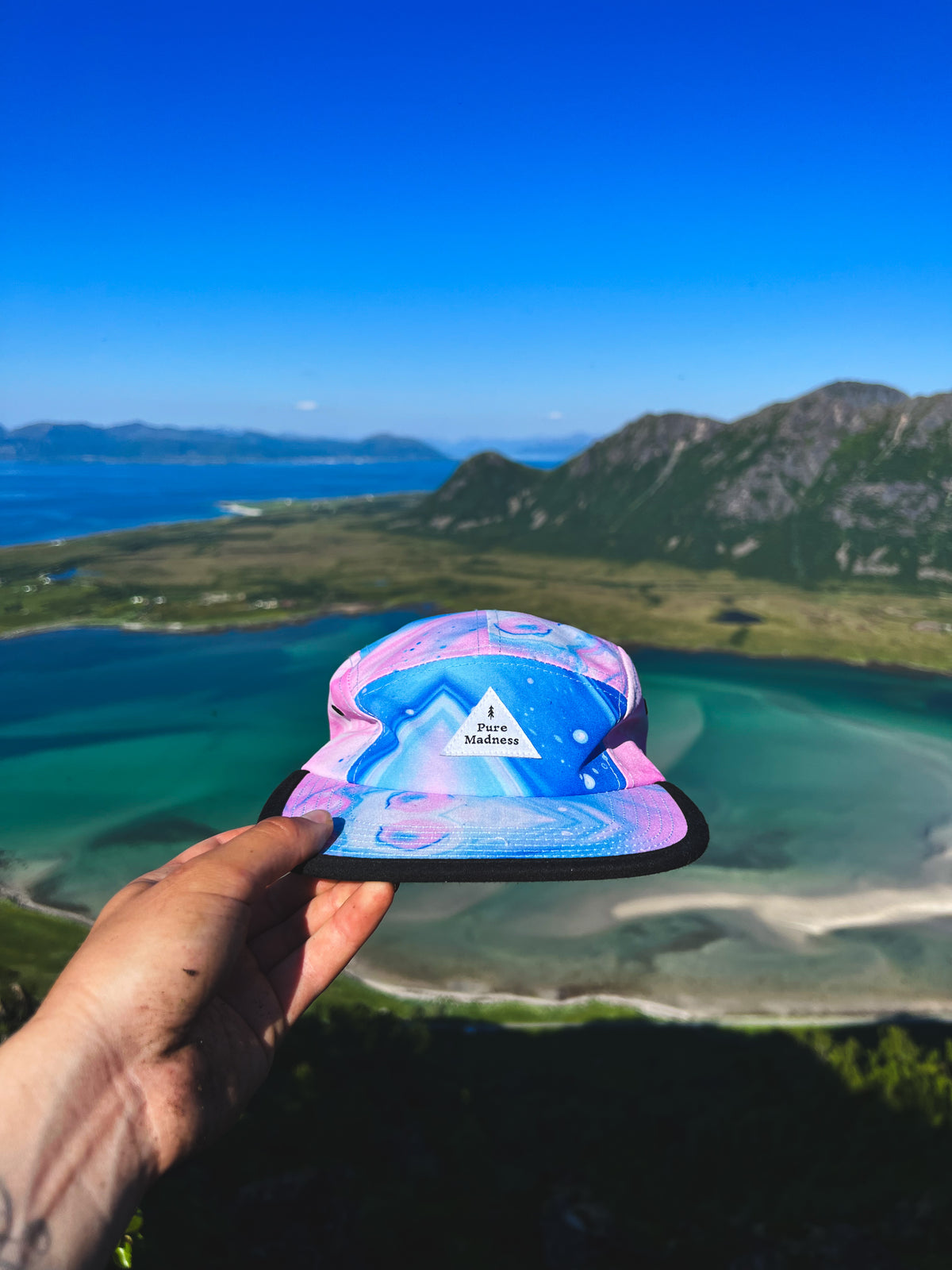 5 Panel Cap Vosso – caps i blått og rosa marmormønster holdt opp over fjord og strand