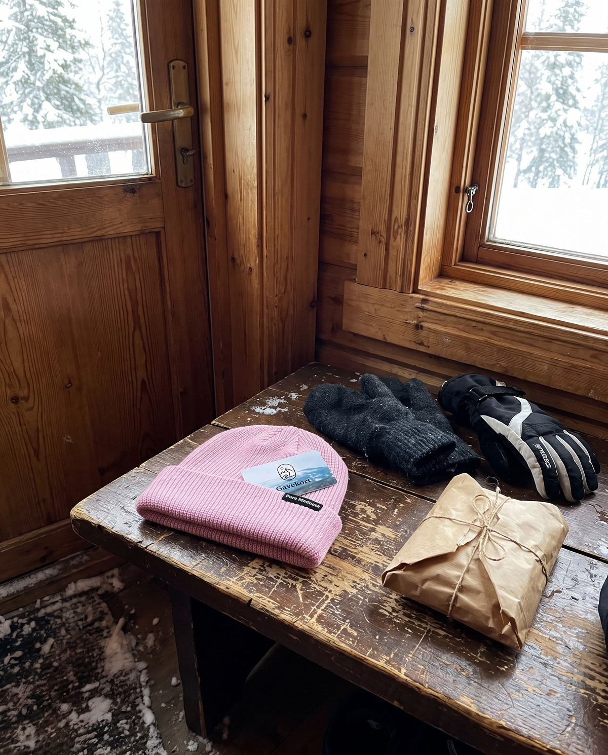 Gavekort – rosa lue, votter og en innpakket gave på et bord i en hytte