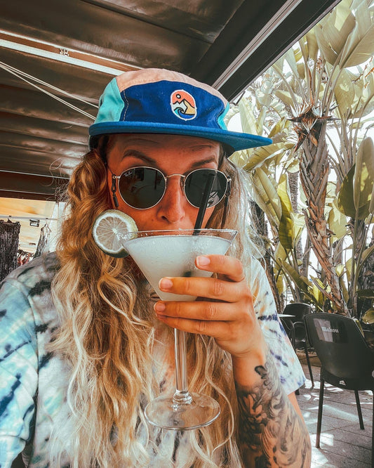5 Panel Cap Ocean Sunset – kvinne med solbriller og capsen med en drink