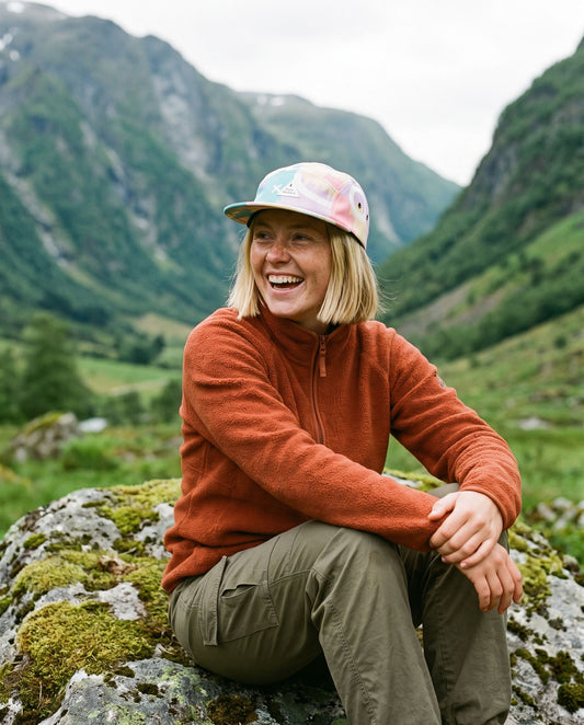 5 Panel Cap Legend – smilende kvinne i oransje fleece sitter på en stein i en grønn fjelldal