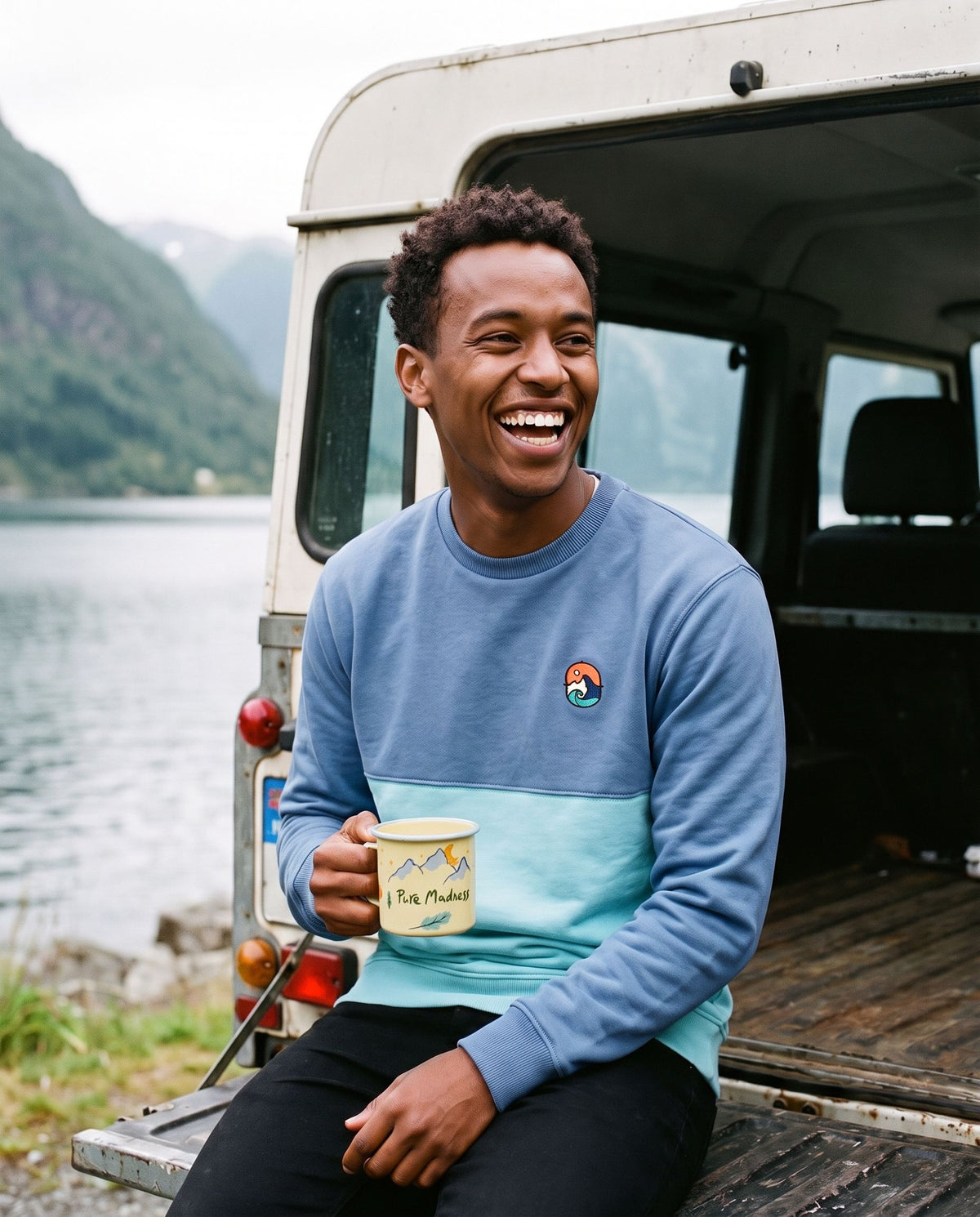 Blue Wave Crew Sweatshirt – leende mann med emaljekrus i bakluka på en jeep ved fjorden