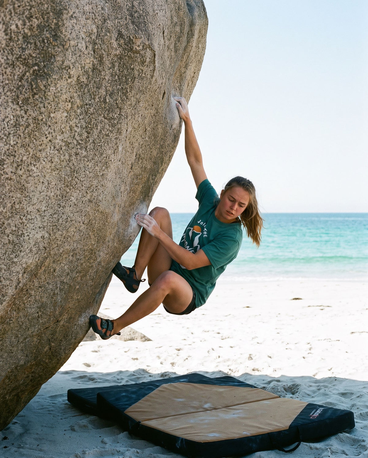 Join the Madness Teal Tee – kvinne buldrer på en stor stein på en strand i Lofoten