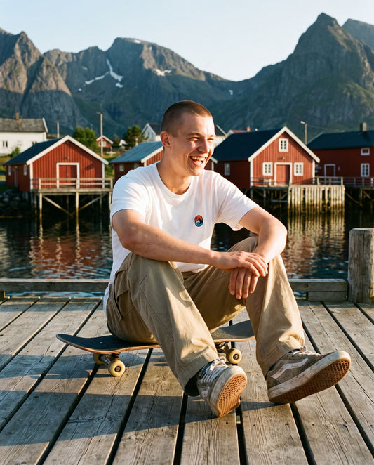 Let the Adventure Begin White Tee – smilende mann med skateboard på brygga i kveldssol i Lofoten