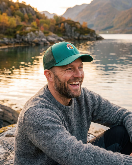 Trucker Cap Teal – smilende mann med capsen og grå ullgenser ved fjorden i kveldssol