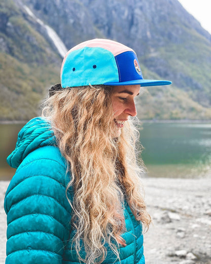 5 Panel Cap Ocean Sunset – kvinne i turkis dunjakke med capsen ved vannet