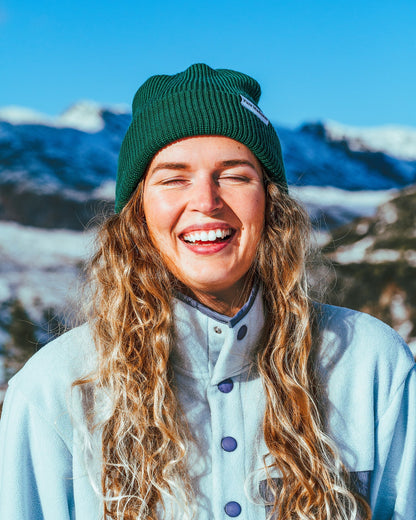 Green Captain Merino Beanie – leende kvinne med grønn lue foran snødekte fjell