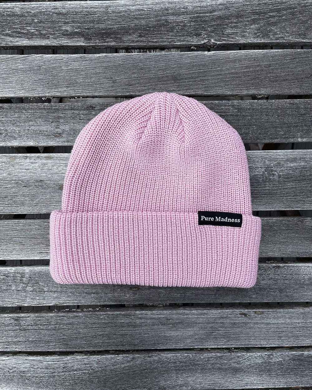 Pink Captain Merino Beanie – rosa merinoull-lue med Pure Madness-merke på grå treplanker
